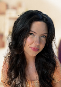 Olivia Smith | Family Guidance & Therapy Center Close-up portrait of a woman with long dark hair and pink lipstick, looking at the camera outdoors with soft lighting.